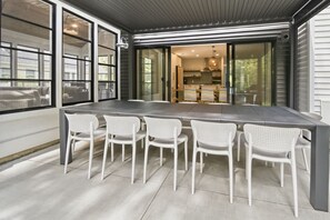 Ein moderner Essbereich mit einem großen Tisch, sechs weißen Stühlen, großen Fenstern und Blick zur Küche.