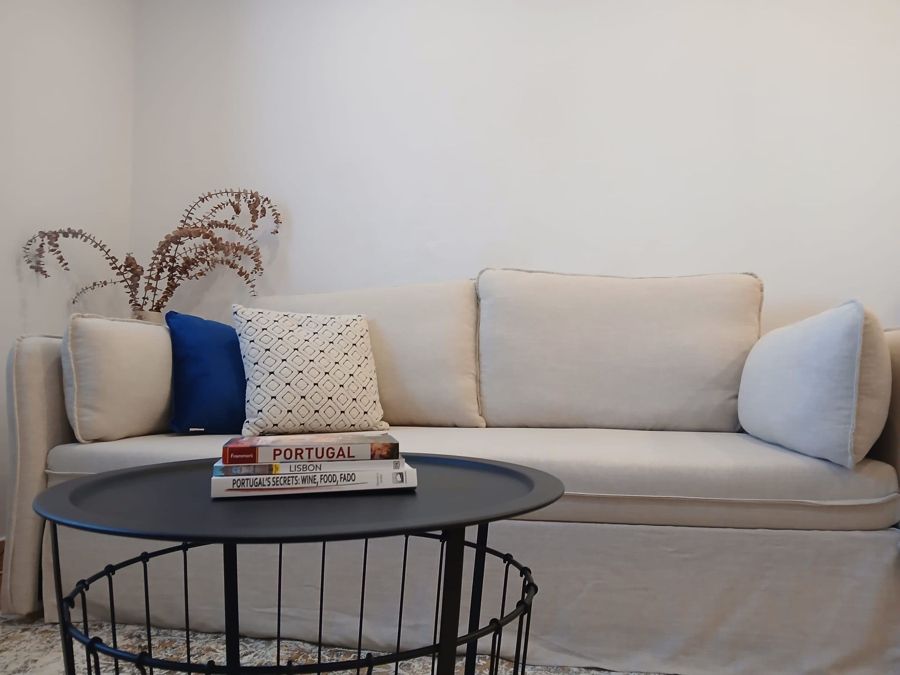 A beige sofa with patterned cushions, a small round table with books, and a decorative plant.
