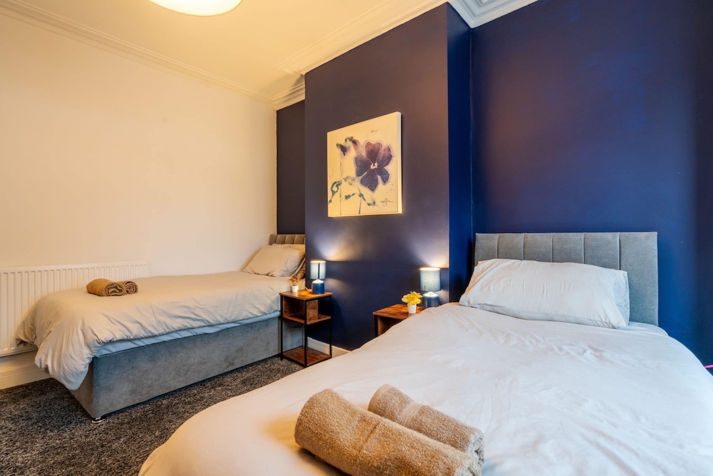 A hotel room with two beds, a nightstand with a lamp, a framed artwork on the wall, and a radiator.
