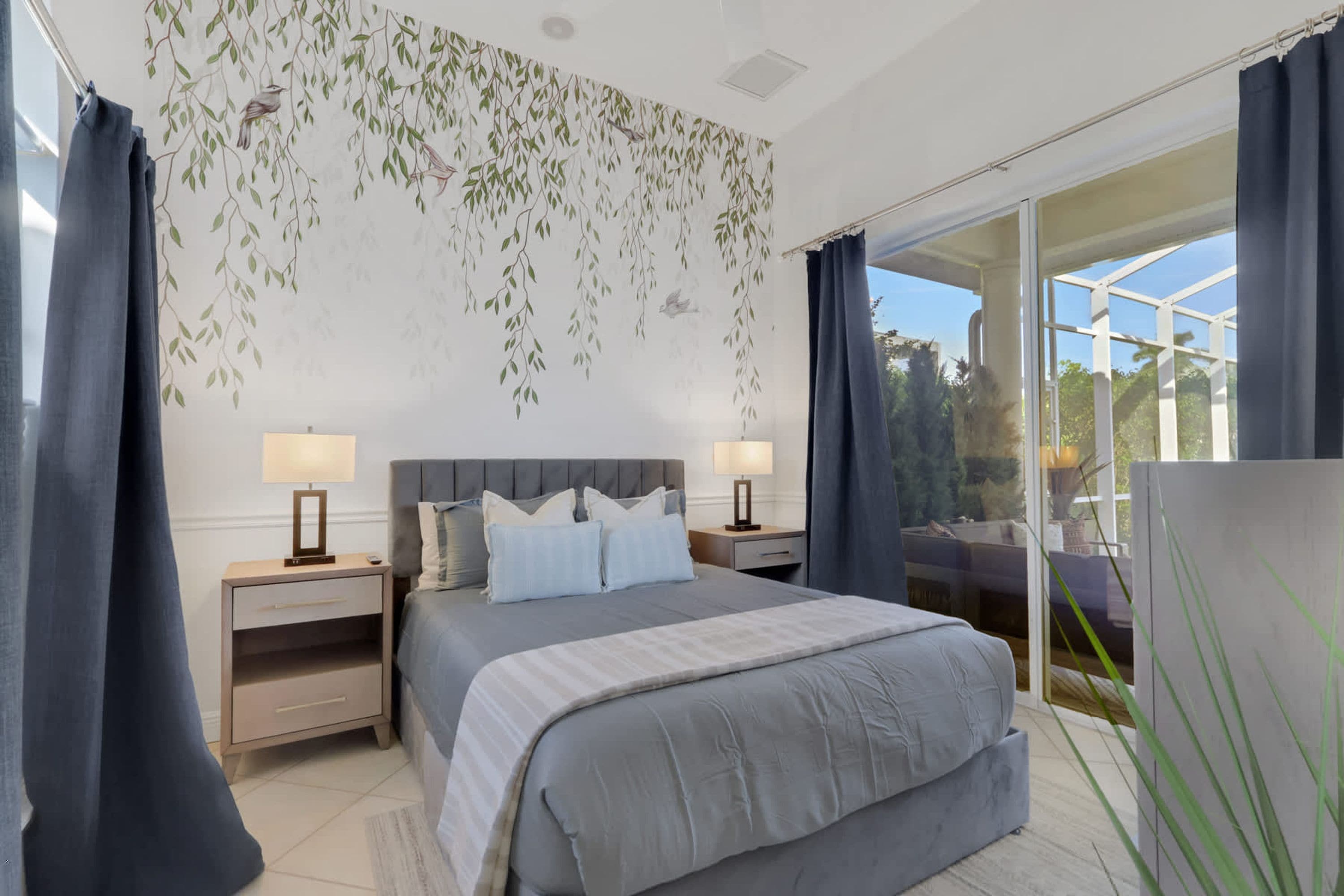 A bedroom with a bed, two bedside tables with lamps, a large window with curtains, and a view of greenery outside.