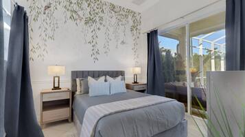 A bedroom with a bed, two bedside tables with lamps, a large window with curtains, and a view of greenery outside.