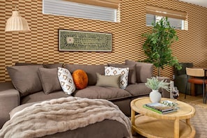 A modern living room with a brown sofa, patterned cushions, a fluffy throw blanket, a round coffee table with books and a vase, and a large plant. - Eastside ATX Social Golf Sim Hot Tub Sauna (Austin)