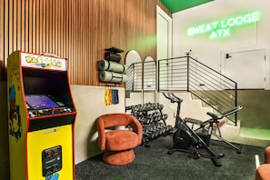A room with a Pac-Man arcade machine, a treadmill, dumbbells, and a neon sign reading 'Sweat Lodge ATX'. - Eastside ATX Social Golf Sim Hot Tub Sauna (Austin)