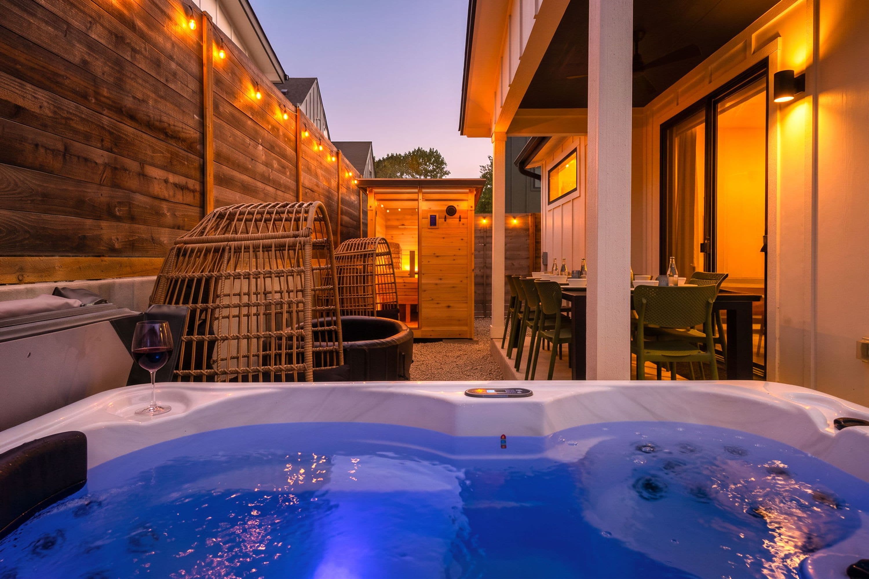A hot tub with wicker chairs and a table, a wooden deck, and a building with a door and windows.