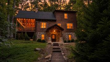 A two-story log cabin with a stone foundation, a porch with string lights, and a stone pathway leading to the entrance.