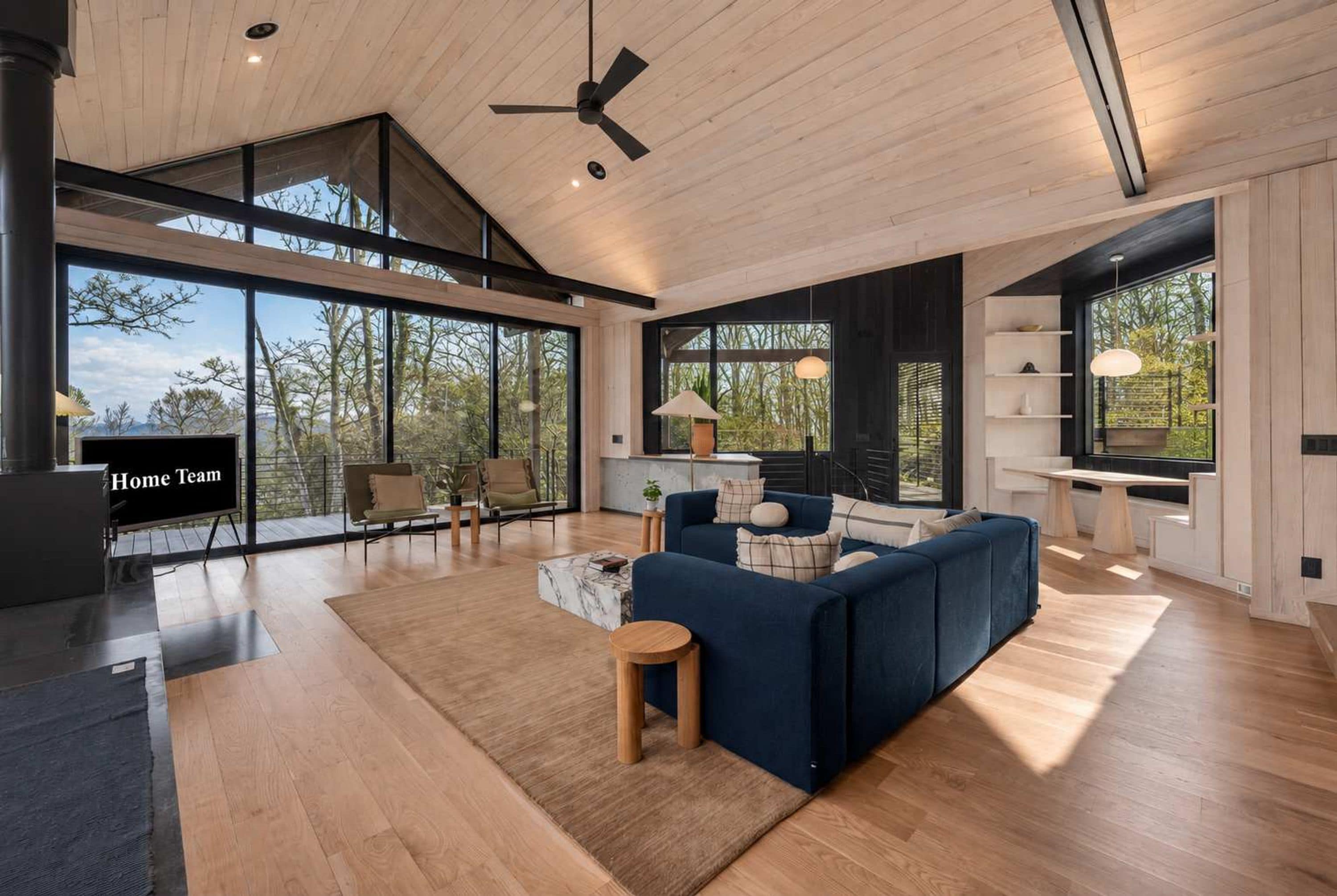 Un amplio salón con un sofá grande, una mesa de centro y un televisor. Grandes ventanales ofrecen vistas al exterior.