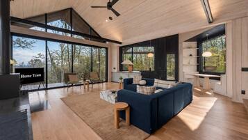 Un amplio salón con un sofá grande, una mesa de centro y un televisor. Grandes ventanales ofrecen vistas al exterior.
