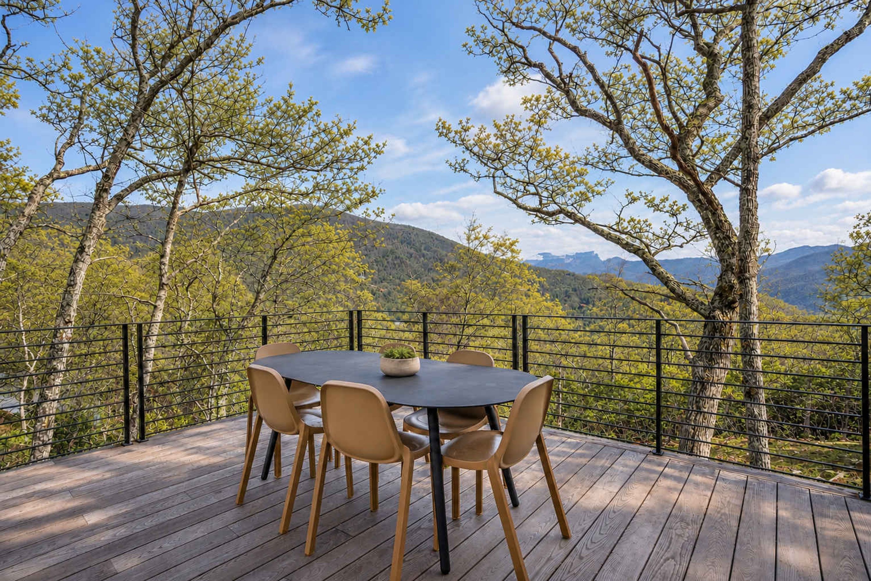 Un área de comedor al aire libre con una mesa y sillas, rodeada por una barandilla, con vistas a un paisaje montañoso.