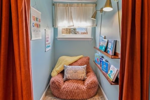 Un coin lecture confortable avec un fauteuil moelleux, une fenêtre avec des rideaux, une étagère fixée au mur remplie de livres et une affiche motivante.