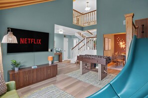 A spacious living room with a large flat-screen TV, a foosball table, a slide, and a staircase leading to an upper level. - 4BR Emerald Coast Escape Pool Hot Tub Theater (Fort Walton Beach)