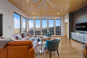 A spacious living room with a large window, leather sofa, and a television on a cabinet. - Blissful Bluff Mtn Views Fire Pit Sleeps 6 (Candler)