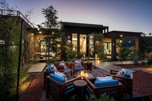 An outdoor seating area with wooden furniture and a fire pit, surrounded by a landscaped garden and a modern building with large windows. - Bell Rock Hacienda Rooftop Deck Hot tub Sauna (Sedona)