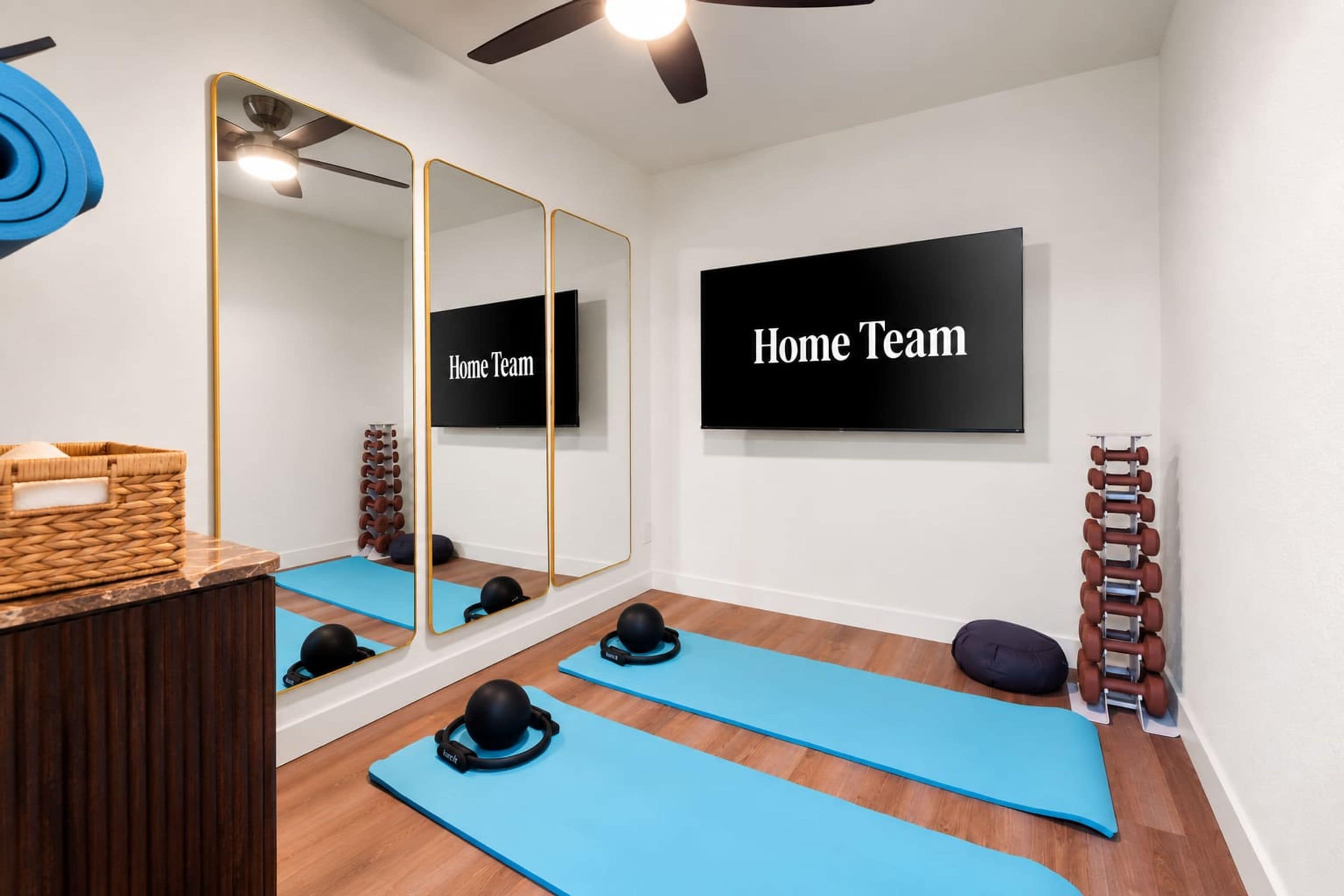Un gimnasio con colchonetas de ejercicio, un espejo, un televisor montado en la pared y un ventilador de techo.
