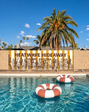 A pool area with a mural of cacti and desert plants, two lifebuoys, and a palm tree in the background. - Heatwave Hideaway Pool Pickleball Games (Tempe)