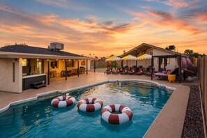 A swimming pool with inflatable rings, a covered patio area, and a sunset sky. - Heatwave Hideaway Pool Pickleball Games (Tempe)