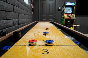 A shuffleboard table with numbered spots, a pool table, and a video game machine in a room with brick walls. - Heatwave Hideaway Pool Pickleball Games (Tempe)