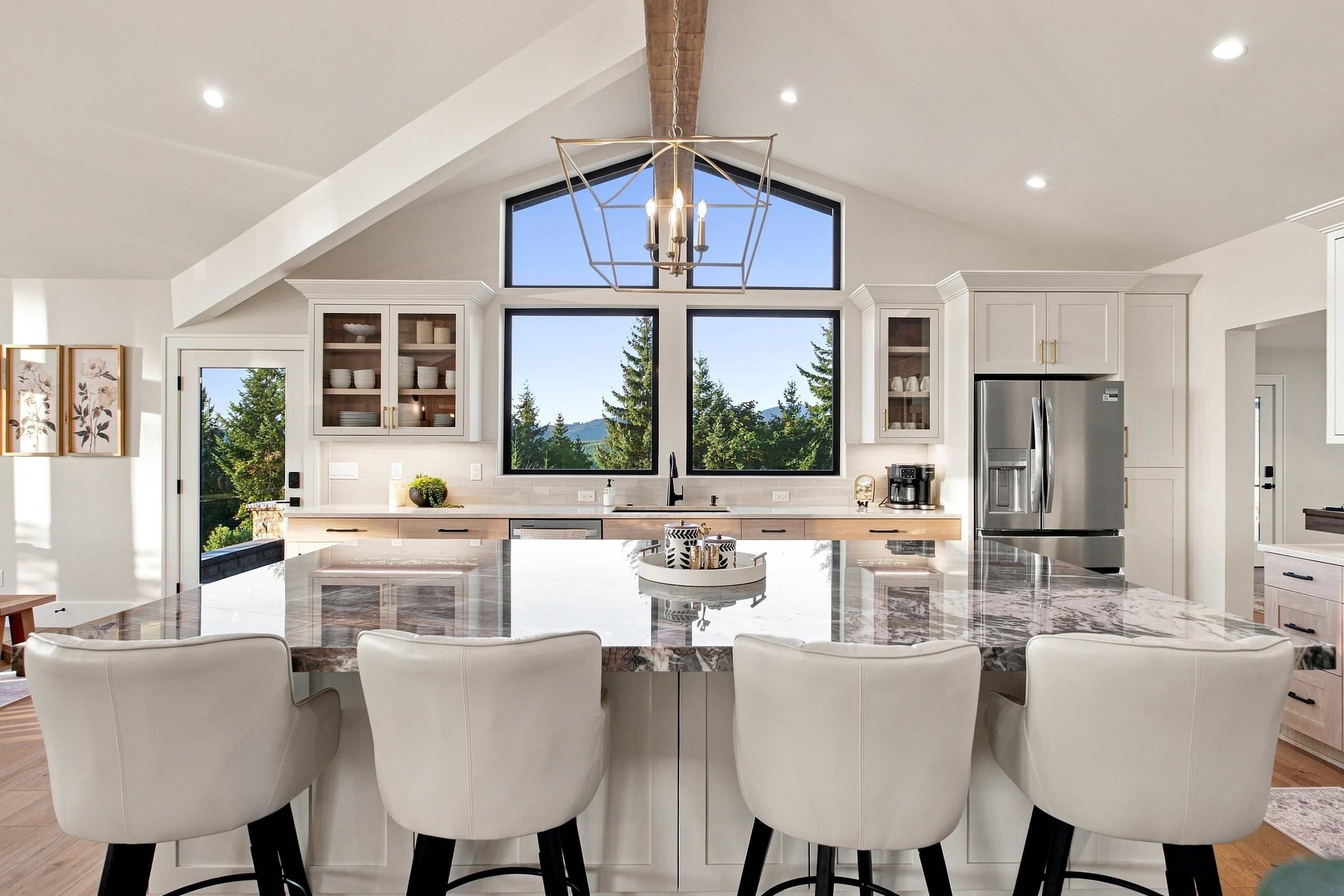 A modern kitchen with a large island, bar seating, and a view of trees through large windows.