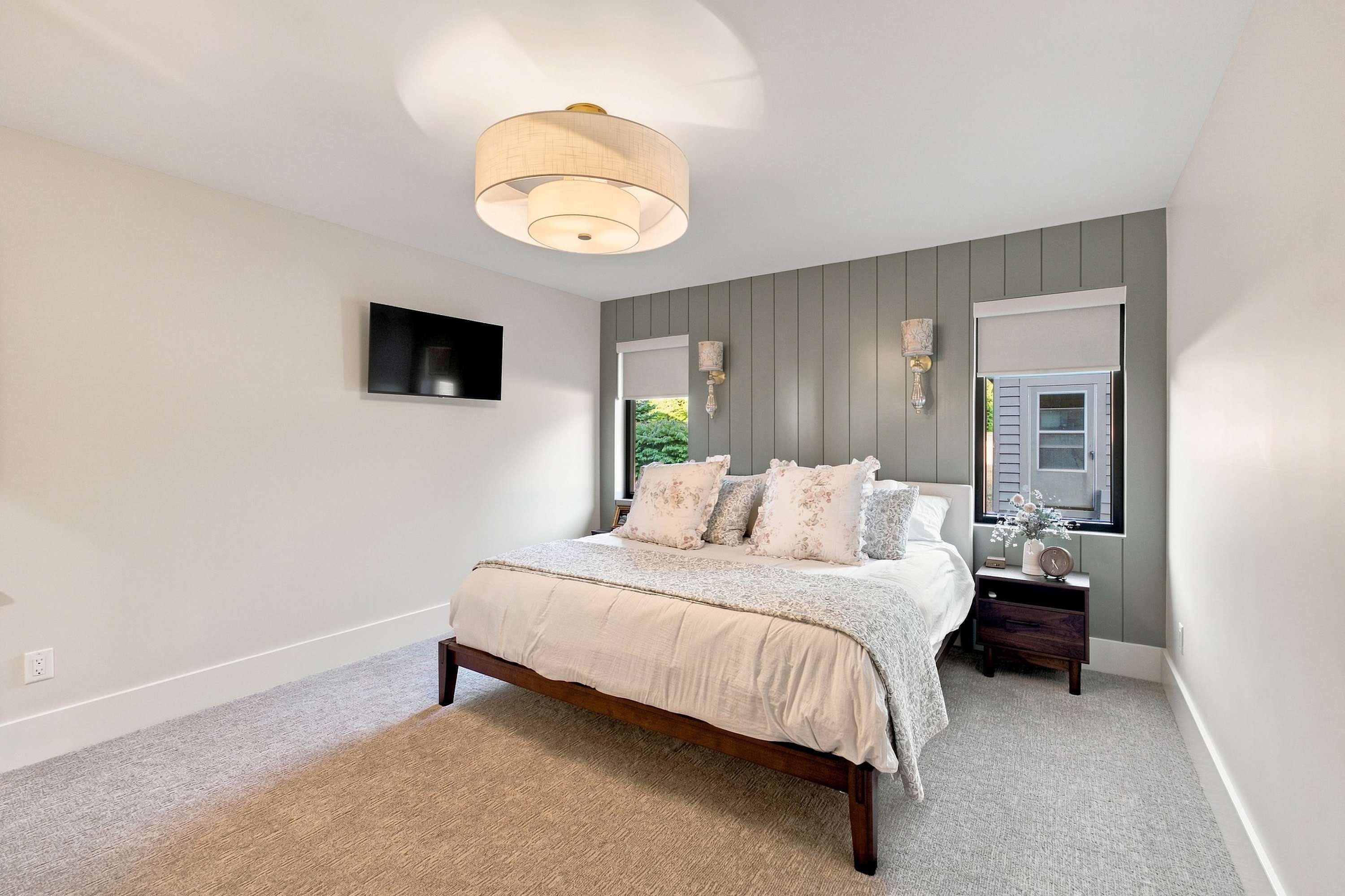 A bedroom with a bed, a nightstand, a wall-mounted TV, and a window with a view of greenery.