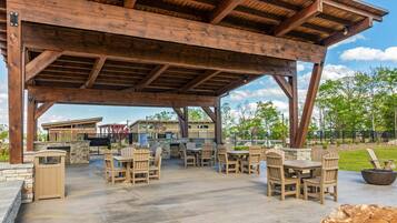 An outdoor patio with wooden tables and chairs, a stone fireplace, and a view of a residential area.