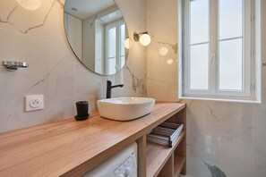 Une salle de bain avec un mur en marbre, un miroir rond, un lavabo blanc et une fenêtre.