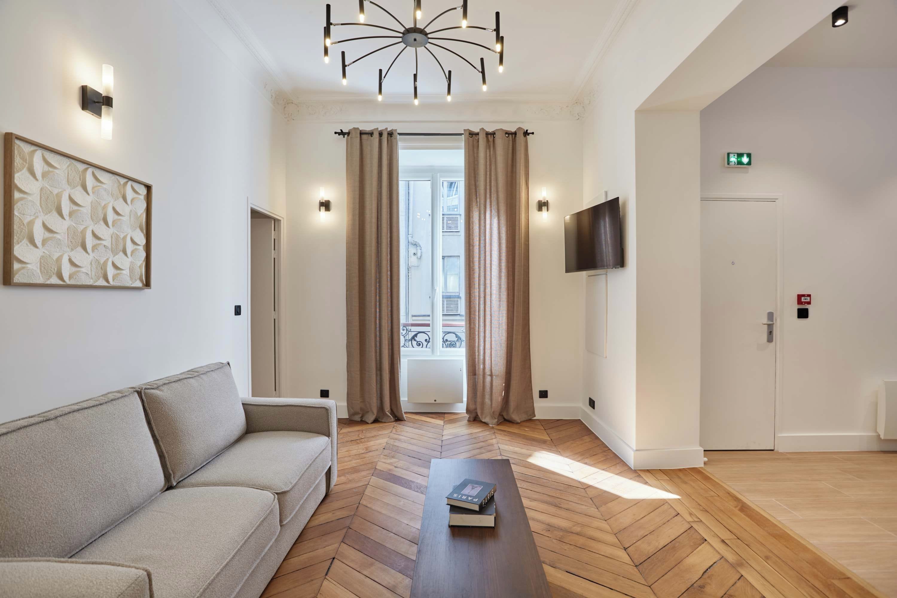 A hotel room with a sofa, a wooden herringbone floor, a flat-screen TV, and a door with an emergency exit sign.