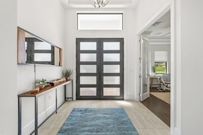 A hallway with a dark wooden door, a light-colored floor, a blue rug, and a large mirror on the wall. - Luxe Waterfront 4 Bed Escape | Near Beach (Marco Island)