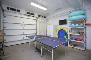 Ein Pingpong-Tisch in einer Garage mit einer weißen Tür, Regalen voller Sportausrüstung und einem blauen aufblasbaren Pool.