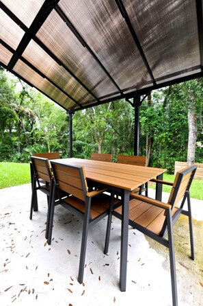 Un patio al aire libre con mesa y sillas de madera, techo metálico y follaje circundante.