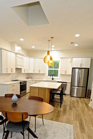 A modern kitchen with white cabinetry, a central island, a stainless steel refrigerator, and a dining area with a wooden table and chairs. - Downtown Bungalow ~ 3BR 3.5BA Home, New Build, Private Yard (Fernandina Beach)