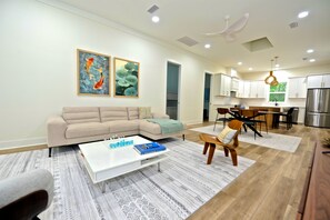 A modern living room with a sectional sofa, a coffee table with books, and a dining area with a wooden table and chairs. - Downtown Bungalow ~ 3BR 3.5BA Home, New Build, Private Yard (Fernandina Beach)