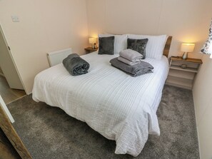 A neatly made bed with a white quilt, two pillows, and a folded gray towel. There are two bedside tables with lamps and a small shelf. - Frontline Sea View : P1 Whilk Meadow Caravan Park (Girvan)