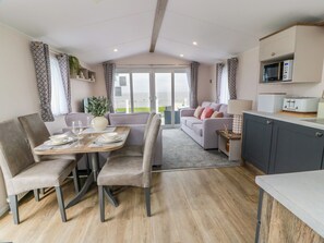 A modern living room with a dining area, a sofa, a kitchen with a microwave, and a view through the glass doors. - Frontline Sea View : P1 Whilk Meadow Caravan Park (Girvan)