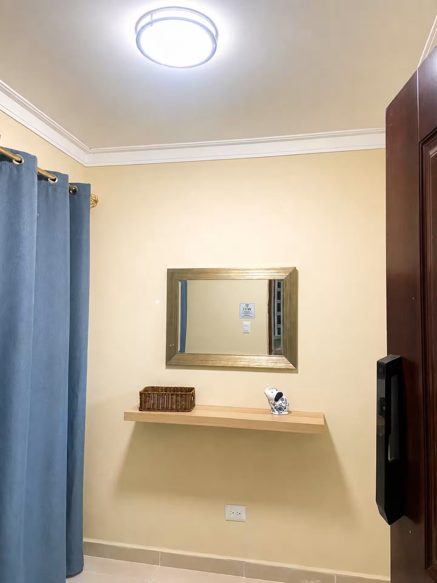 A bathroom with a ceiling light, a mirror, a shelf with a basket and a decorative item, and a door.