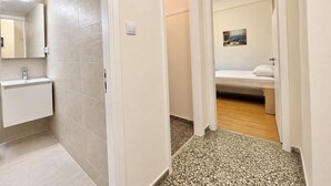 Corredor que dá para um banheiro com piso ladrilhado, pia e cama com cabeceira branca.