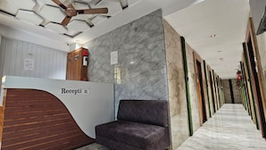 A reception area with a check-in/out sign, a reception desk, a seating area, and a hallway with doors. - Hotel Alfa View (Ahmedabad)
