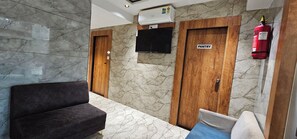 A lobby with a sofa, a flat-screen TV mounted on the wall, wooden doors, and a fire extinguisher.