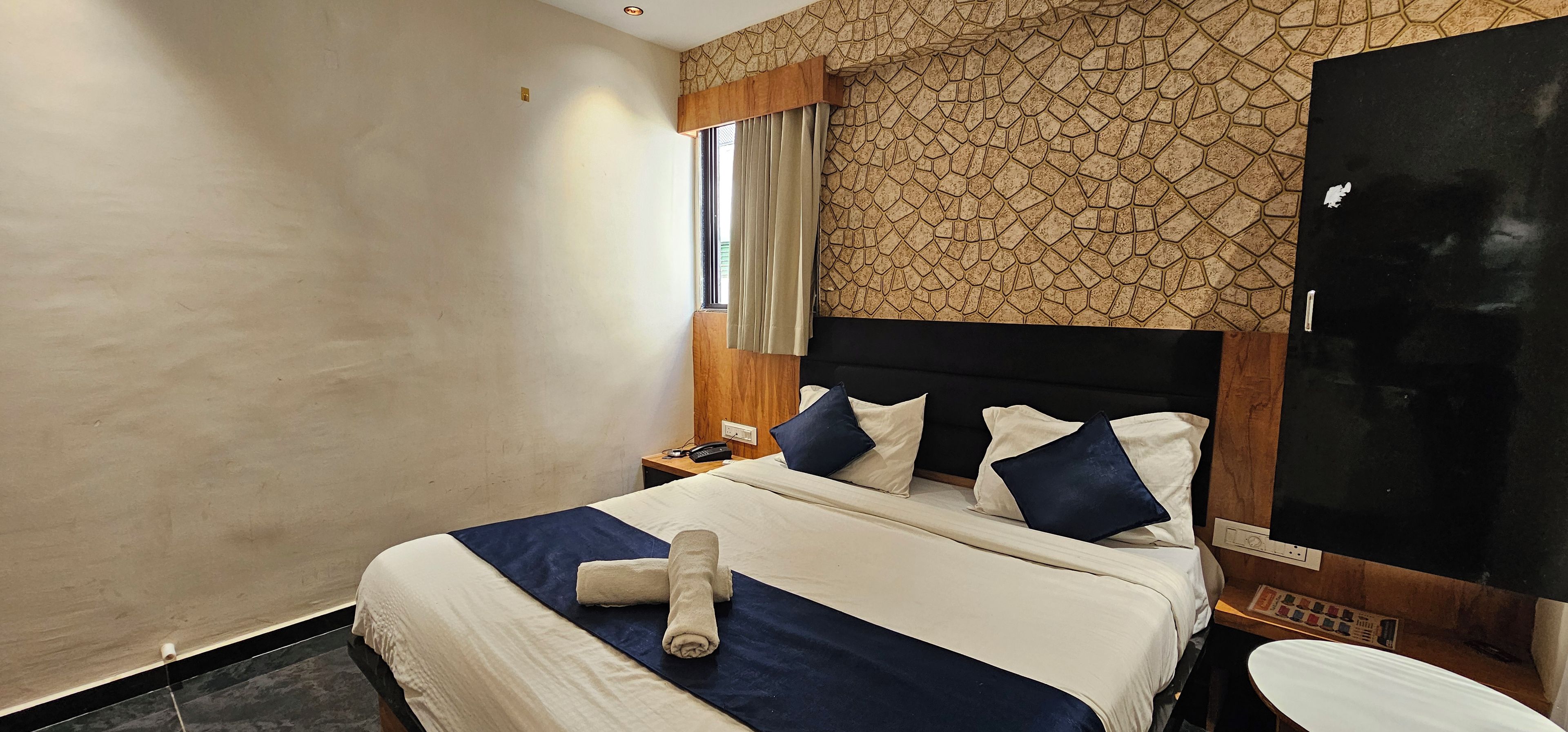 Quarto de hotel com cama, mesa de cabeceira, TV fixada na parede e janela com cortinas.