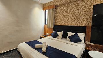 Quarto de hotel com cama, mesa de cabeceira, TV fixada na parede e janela com cortinas.