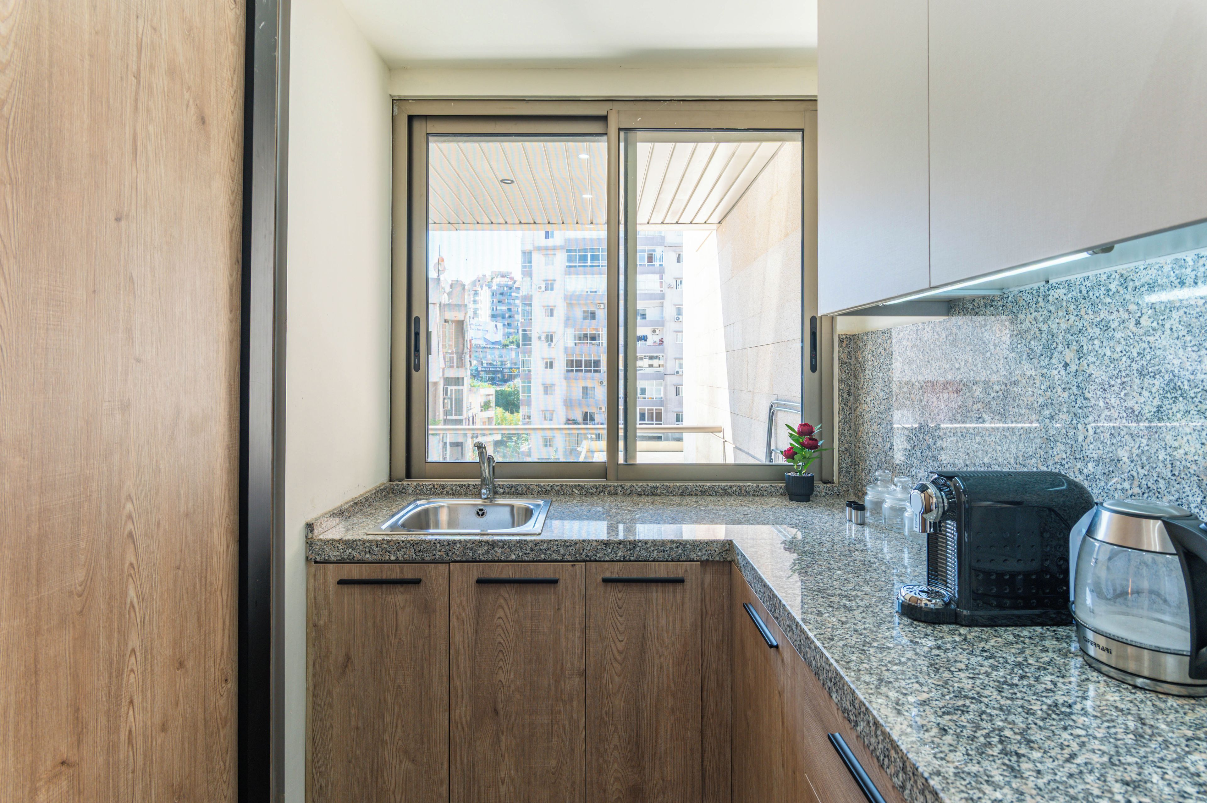 Eine moderne Küche mit Spüle, Holzschränken, einer Arbeitsplatte aus Granit und Blick auf eine Stadtlandschaft durch ein großes Fenster.