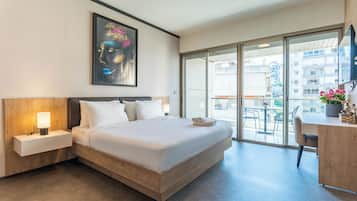 A hotel room with a large bed, a bedside table with a lamp, a painting on the wall, and a view of the city through the balcony doors.