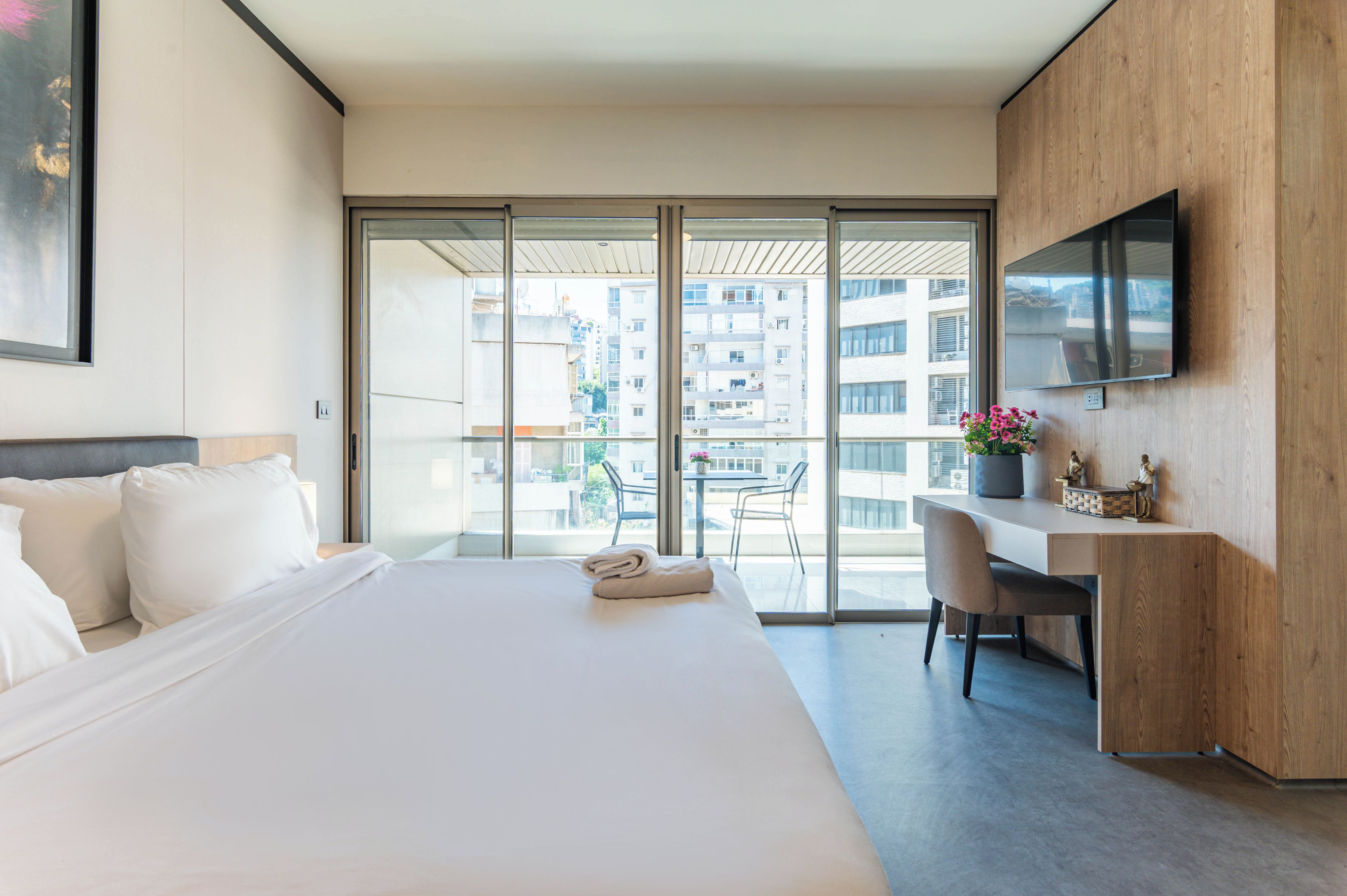 A modern hotel room with a large bed, a desk, a chair, a TV, and a balcony with a view of the city.