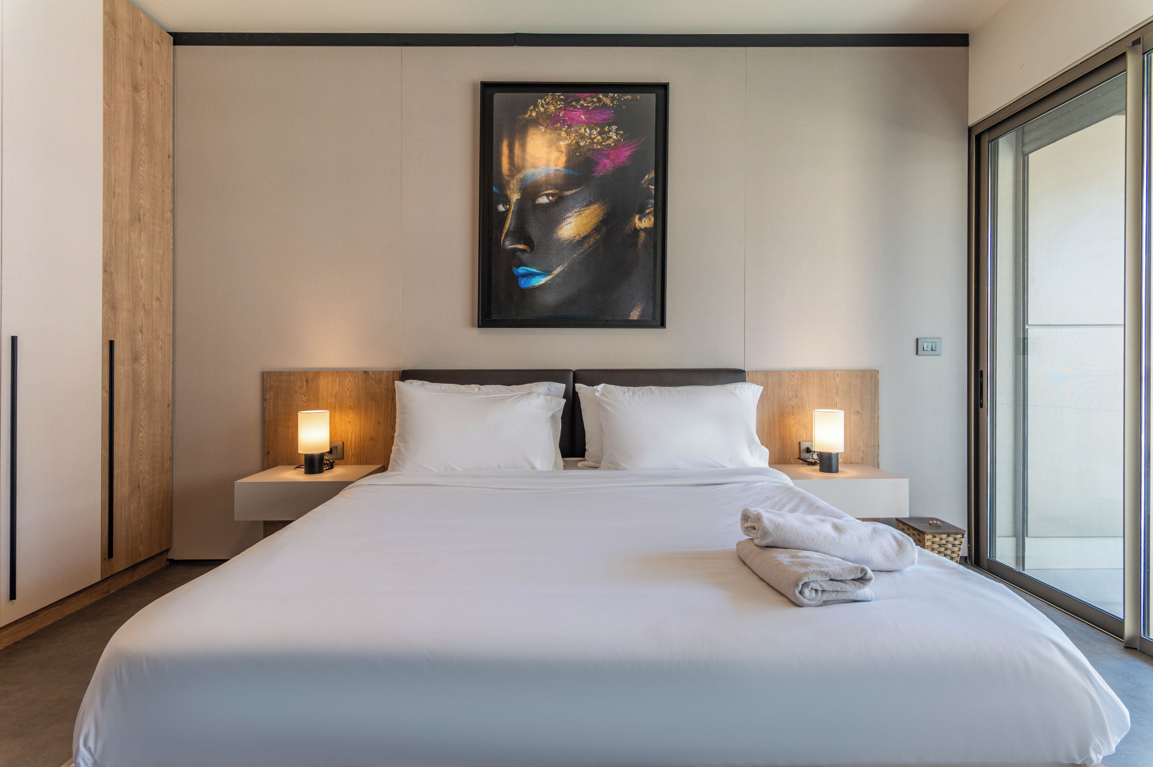 Habitación de hotel con una cama grande, dos mesitas de noche con lámparas, un ventanal amplio y un cuadro en la pared.