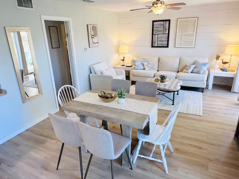 Una sala de estar con un juego de sofá, una mesa de comedor con sillas, un ventilador de techo y decoración en la pared.
