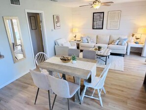 Una sala de estar con un juego de sofá, una mesa de comedor con sillas, un ventilador de techo y decoración en la pared.