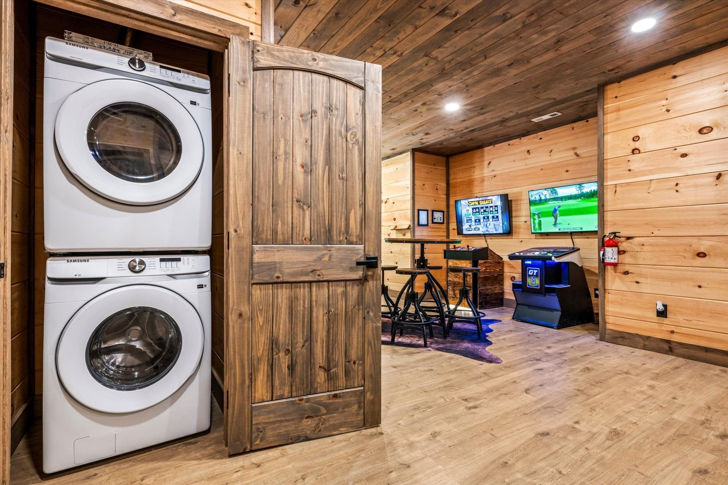 A laundry room with two Samsung washing machines, wooden walls, and a gaming area with a pool table and two TVs.
