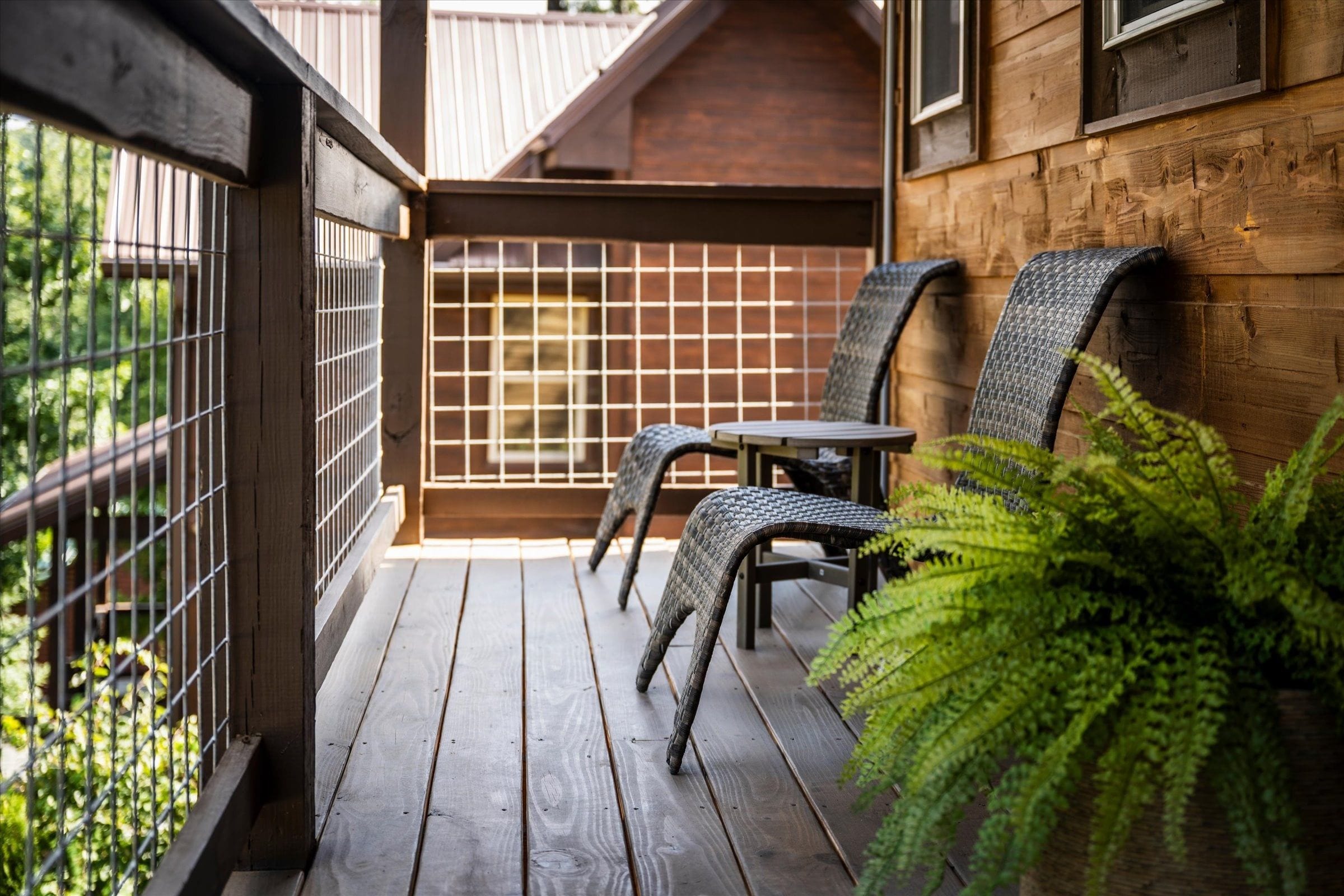 A wooden balcony with a metal fence, two chairs, a small table, and a potted plant.