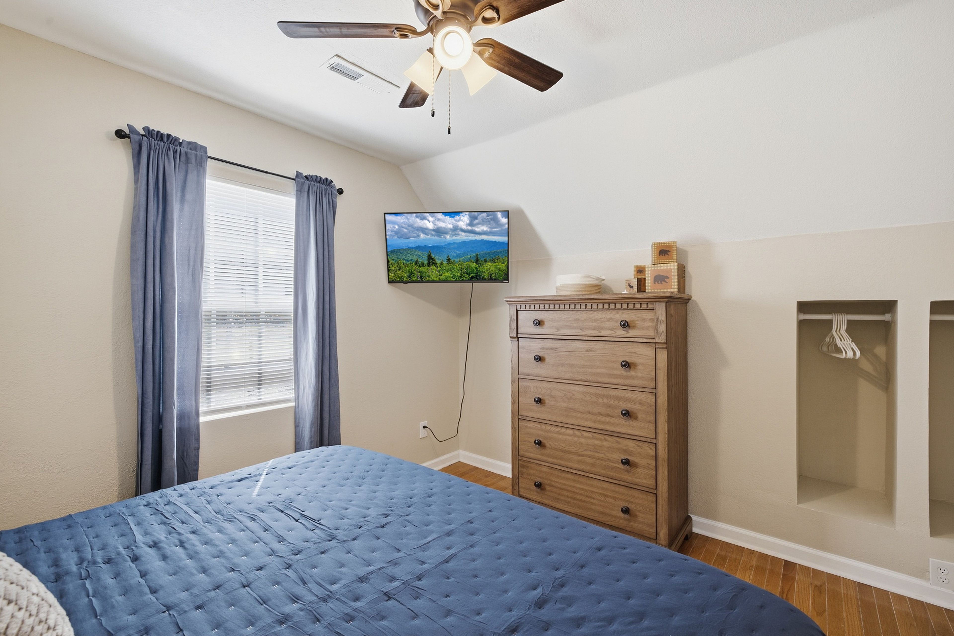 A bedroom with a bed, a dresser, a television mounted on the wall, and a ceiling fan.