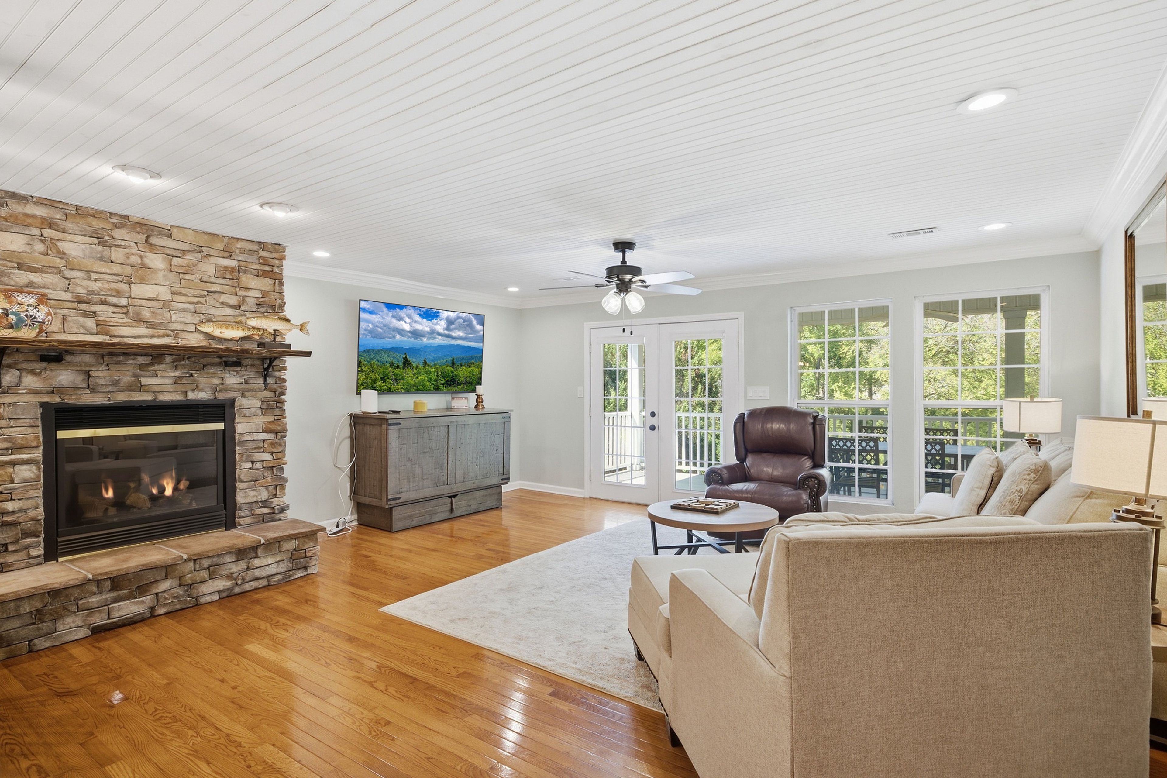 A living room with a stone fireplace, a leather armchair, a sofa, and a television on the wall.