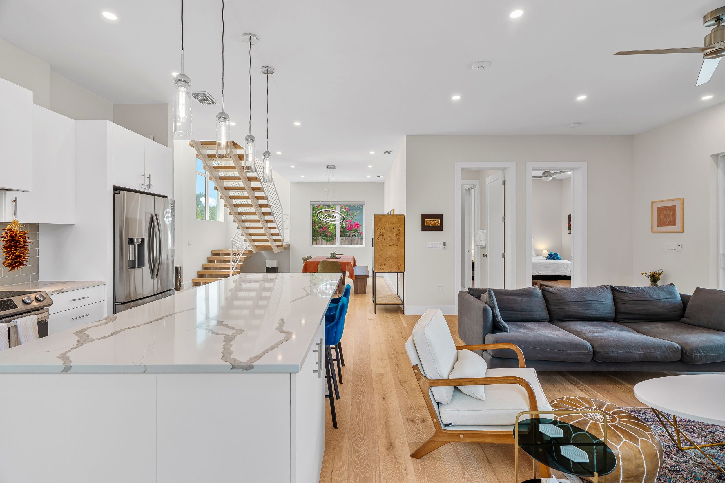 A modern kitchen with a central island, a dining area with a wooden table, a grey sofa, and a staircase leading to an upper level.