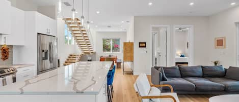 A modern kitchen with a central island, a dining area with a wooden table, a grey sofa, and a staircase leading to an upper level.